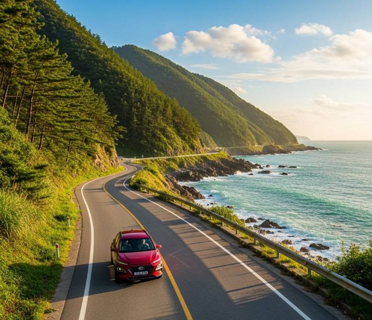 A scenic, winding coastal road on Namhae Island, South Korea, with a car driving alongside a dramatic ocean view and green hills.