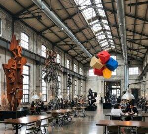 The vast, open interior of Daerim Changgo, a factory gallery cafe in Seoul, showing high ceilings, exposed brick walls, and large contemporary art sculptures amidst dining tables