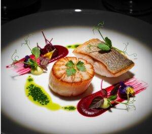 An elegantly plated fine-dining dish featuring a seared scallop with artistic sauce and microgreens on a white plate.
