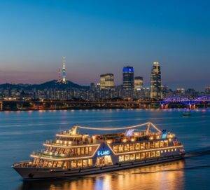 The E-land cruise ship, fully illuminated, sailing on the Han River at night with the Seoul skyline in the background.