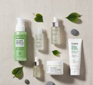A flat lay of several Korean skincare products specifically for acne. The products include a cleanser, toner, essence, and moisturizer, arranged neatly on a minimalist, light-colored background with some green botanical elements.