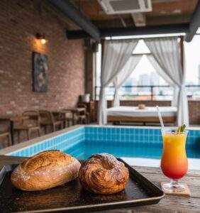 Cafes in Mullae Art Village: A close-up, food-blogger style shot of a rustic sourdough loaf and a glistening cruffin on a metal tray, with the blurred industrial-chic interior of a bakery (red brick walls and exposed air ducts) in the background.
