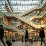 Wide-angle landscape photograph of the bustling multi-level Yongsan I'Park Mall guide: interior at golden hour, featuring crisscrossing escalators under a large glass ceiling and travelers with suitcases walking.