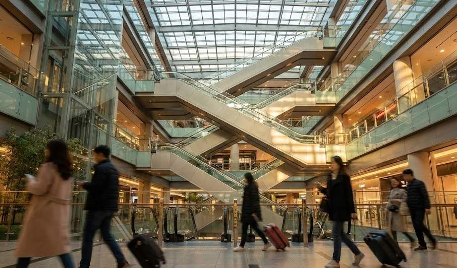 Wide-angle landscape photograph of the bustling multi-level Yongsan I'Park Mall guide: interior at golden hour, featuring crisscrossing escalators under a large glass ceiling and travelers with suitcases walking.