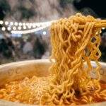 Tasted Real Shin Ramyun: A close-up photograph at night showing chopsticks lifting steaming, curly noodles from a pot of spicy boiling ramen broth, with blurred festival lights and crowds in the background.