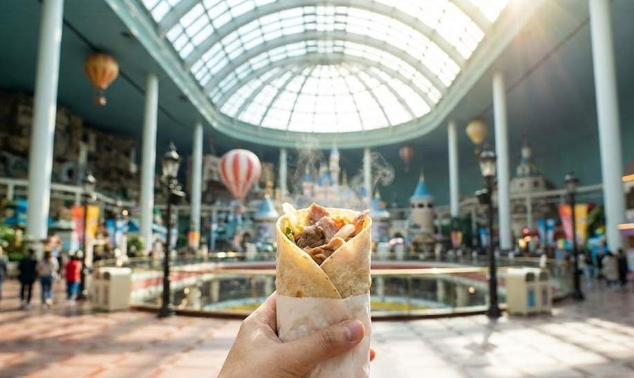 Lotte World Halal Guide: A first-person view holding a delicious Halal chicken kebab wrap inside the bustling Lotte World Adventure indoor theme park under the glass dome ceiling.