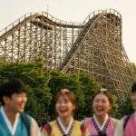A scenic view of Everland's T-Express wooden rollercoaster at sunset, with young tourists in rented Korean school uniforms laughing in the foreground.