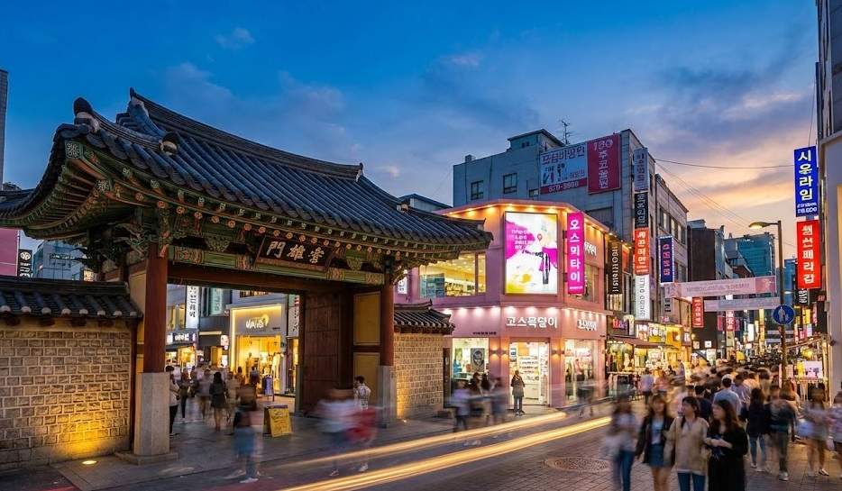 A bustling street scene in Myeongdong, Seoul, at twilight, showing a traditional Korean gate (Heunginjimun style) next to modern buildings with glowing neon signs in Hangul. People are walking with motion blur.