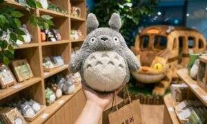 First-person point-of-view photograph of a hand holding a grey Totoro plush toy inside the Ghibli store. A large Catbus photo spot is visible in the warm-lit background filled with wooden shelves of merchandise.