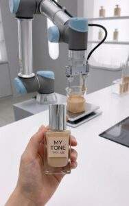 First-person point of view holding a custom "My Tone" foundation bottle at the Amore Store's Base Picker lab, with a robotic arm mixing foundation in the background.