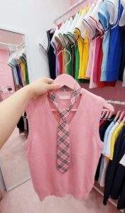 First-person view of a hand holding a pink knit vest and plaid tie in a Korean school uniform rental shop.