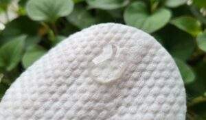 A macro photograph of the embossed side of an Anua Clear Pad, showing its bumpy texture. A clear drop of liquid essence glistens on top of the pad. The background is blurred green heartleaf plants.