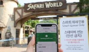 A first-person point of view shot looking down at a smartphone screen showing a confirmed Everland app booking for Safari World. In the background, the entrance archway and a sign reading "Smart Queue Only" are visible.