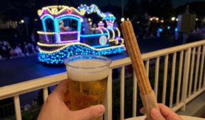 First-person perspective holding a beer and churro on a balcony railing, with a bright LED parade float passing in the background at Everland.