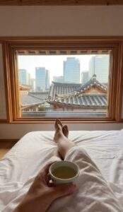 A vertical first-person POV shot from a comfortable bed in a modern Hanok hotel room, holding a cup of tea and looking out a low wooden window framing traditional Korean tiled roofs and modern skyscrapers.