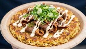 Close-up food photograph of a crispy ramen noodle pancake topped with spicy sliced beef, green onions, and garlic mayo sauce on a paper plate.