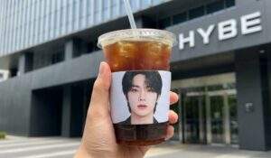 First-person perspective photo of a hand holding an iced americano with a K-Pop idol cup sleeve in front of the HYBE building entrance.