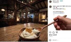 A first-person point of view shot from an Instagram post, showing a hand scooping honeycomb Greek yogurt with a wooden spoon at Dotori cafe. The background is a dimly lit, rustic, Ghibli-themed interior.