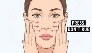An instructional illustration of a woman's face with arrows indicating a pressing motion on the cheeks and text reading "PRESS, DON'T RUB"