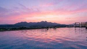 A wide-angle view of people relaxing in the rooftop infinity pool at Aquafield Goyang during a pink and purple sunset, overlooking mountains.