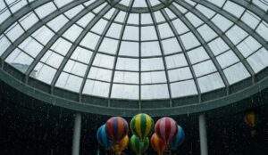 A close-up view looking up at the Lotte World glass dome ceiling, with raindrops streaking across the glass and colorful hot air balloons from a ride floating below.