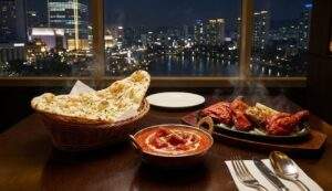 A luxurious spread of Indian food including naan bread, butter chicken curry, and tandoori chicken on a table at Agra restaurant. The window background shows a stunning night view of Seokchon Lake and the Seoul city skyline.