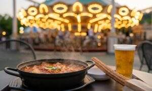 Everland Korea: A close-up photograph of a Korean meal at Everland featuring Kimchi stew, churros, and beer on an outdoor table at twilight with a carousel in the background.