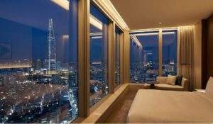 A luxurious hotel room interior in Gangnam at night, featuring floor-to-ceiling windows with a panoramic view of the Seoul skyline and the illuminated Lotte World Tower.
