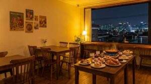 Cozy interior of a Yongridan-gil cafe at night, featuring wooden furniture, pastries, Tteokbokki, and a large window with a view of Seoul's city lights.