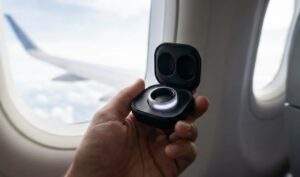 A first-person perspective shot of a hand holding the open Samsung Galaxy Ring charging case with the ring inside, on an airplane with a window view of clouds.