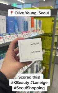A first-person view of a hand holding a Laneige product in an Olive Young store. Social media overlay text reads "📍 Olive Young, Seoul" at the top and "Scored this! #KBeauty #Laneige #SeoulShopping" at the bottom.