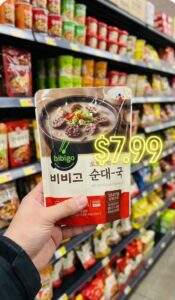 A first-person perspective shot of a hand holding a "Bibigo Sundae-guk" retort pouch in front of a grocery store shelf, likely in an H-Mart. The background shows blurred shelves filled with various Korean food products. A glowing neon-style price tag with the text "$7.99" is floating next to the pouch.