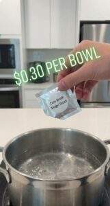 A first-person point-of-view shot of a hand holding a small silver packet labeled "Coin Broth Magic Stock" over a pot of boiling water in a kitchen. The text "$0.30 PER BOWL" is floating above the hand.