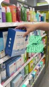 First-person view of a shopper grabbing a blue Round Lab "1+1 Special Set" box from an Olive Young shelf, with neon green 3D text floating nearby reading "$12 EACH".