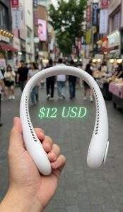 POV holding a white portable neck fan in a busy Myeongdong street with price text $12 USD.