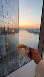 A vertical first-person view from a high-floor luxury hotel room, where a hand holds a coffee cup against a window reflecting a stunning sunset over the Han River and the Seoul cityscape.