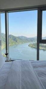 Vertical first-person perspective from a luxury hotel bed, looking out at a panoramic Han River and mountain view with a glass of red wine on the side table.