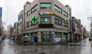 Street view of a typical Seoul neighborhood building on a rainy day, featuring a prominent green cross sign and Hangul text indicating a local internal medicine clinic ("내과").