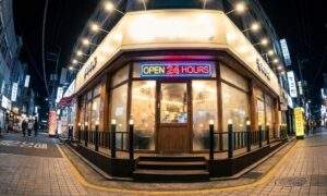 A wide-angle night photograph of a bustling street in Seoul, South Korea, featuring a brightly lit restaurant on a corner. A neon sign above the entrance reads "OPEN 24 HOURS."