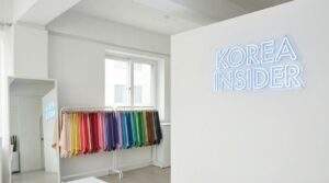 A wide-angle view of a minimalist, modern personal color analysis studio in Seoul. Colorful fabric drapes hang neatly on a rack next to a large mirror. A glowing blue neon sign on the white wall reads "KOREA INSIDER".
