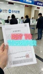 First-person perspective photo inside a Korean bank (Shinhan/Woori). A hand holds a Korean "Tax Payment Certificate" (납세증명서) document. Glowing text overlay says "KEY TO TRANSFER".